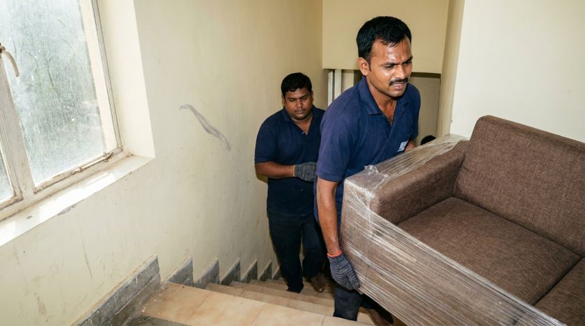 Two moving crew members carrying a wrapped sofa up a narrow staircase in a walk-up apartment building