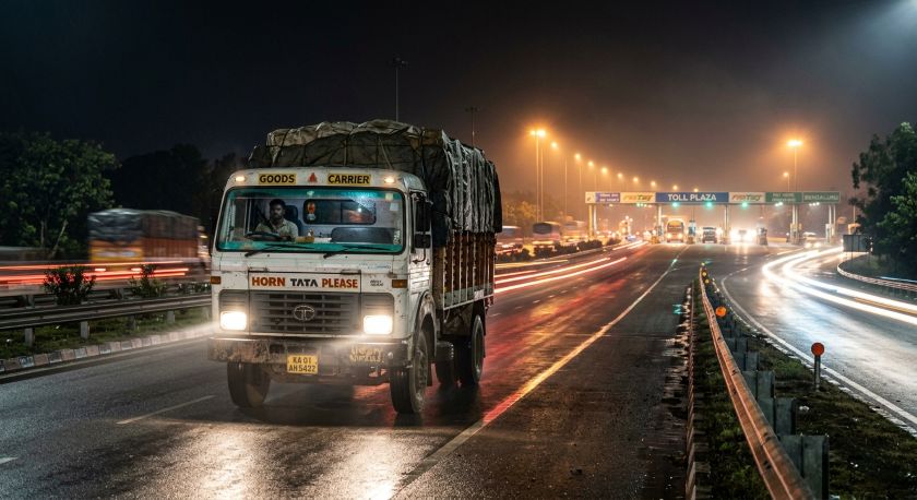 A white fully-loaded 16-ton Indian freight truck with glowing headlights driving on a dark four-lane NH-48 expressway at night, illuminated FASTag toll booth visible 200 metres ahead, light trails on wet tarmac, realistic long-exposure photography style, Nikon D850 wide angle, hyperrealistic