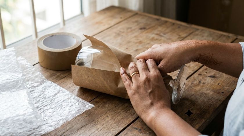Hands wrapping a wine glass stem-first in packing paper before boxing